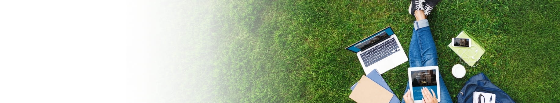 Tablet, laptop, smartphone, notebook, coffee cup, and backpack on grass beside a person lying down.